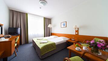 Quarto de hotel com duas camas, uma mesa de escritório com um computador, uma televisão e um espaço de refeições com um vaso de flores.