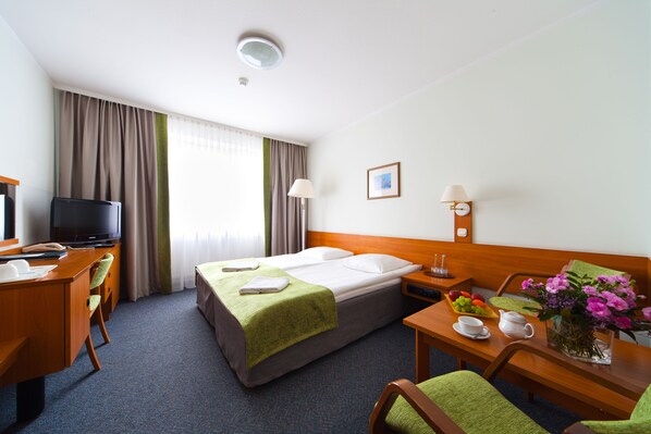 A hotel room with two beds, a desk with a computer, a television, and a dining area with a vase of flowers. - Hotel IOR (Poznan)