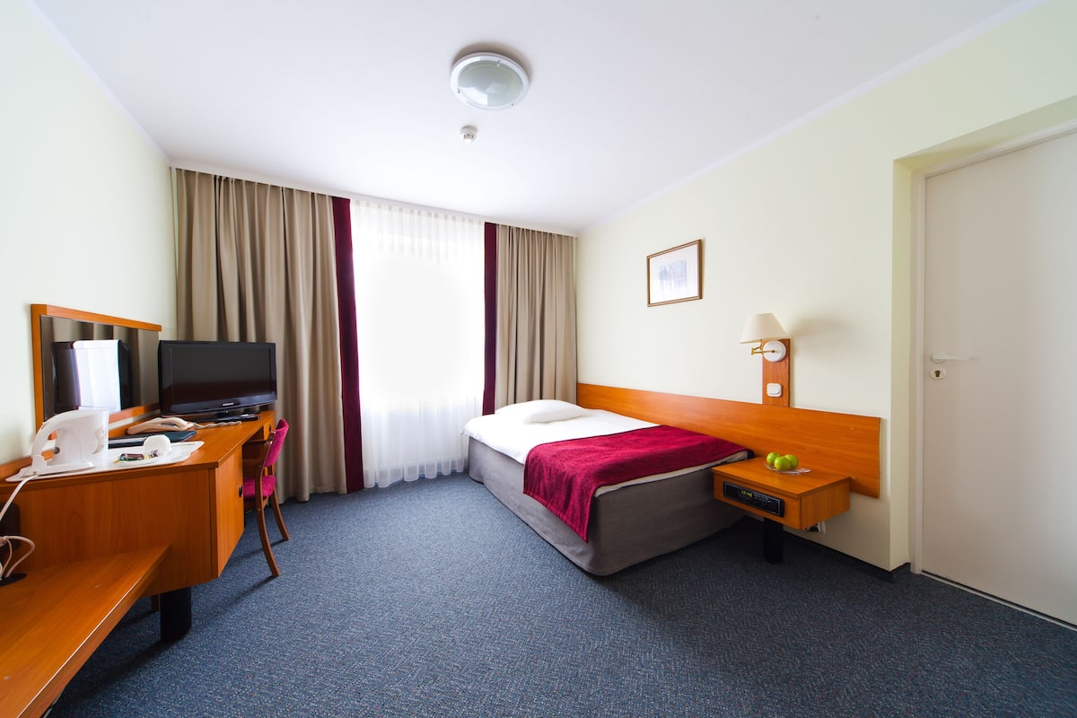 A hotel room with a bed, a desk with a computer, a television, a bedside table with a lamp, and a window with curtains.
