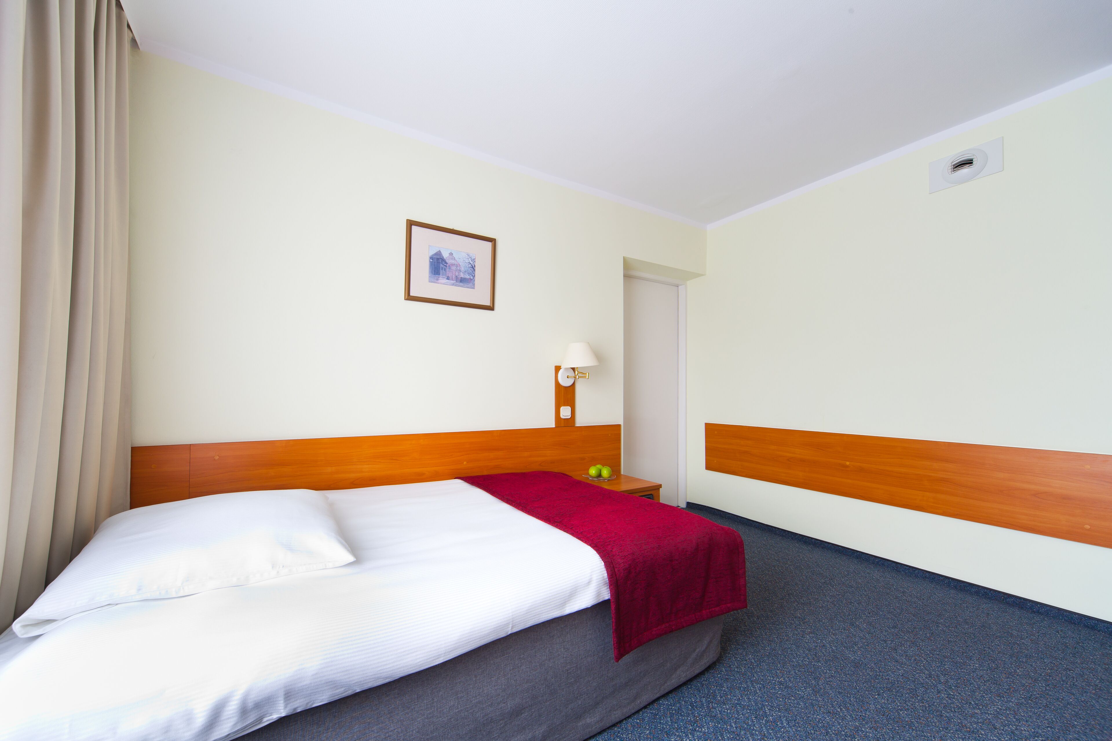 Ein Hotelzimmer mit Bett, Nachttisch, Lampe, einem Bild an der Wand und einer Tür.