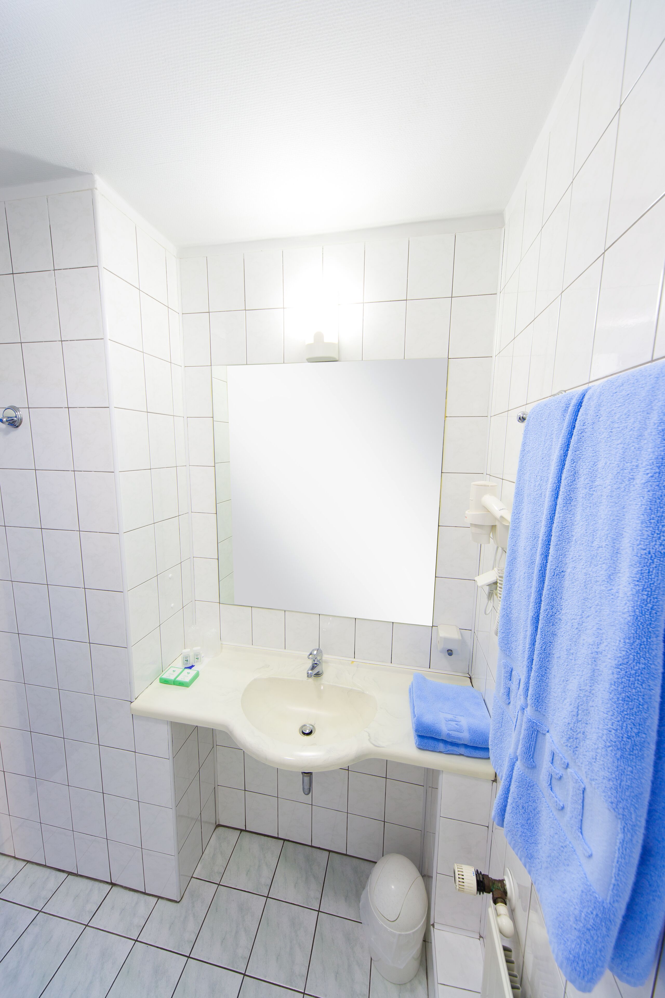 Un bagno con pareti piastrellate di bianco, un lavandino, uno specchio e un asciugamano blu appeso al muro.