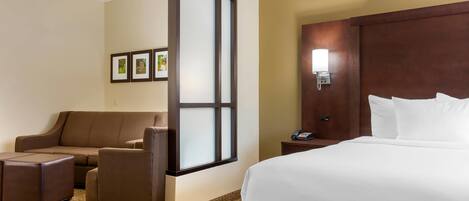 Quarto de hotel com uma cama grande, um sofá marrom, uma poltrona pequena, uma mesinha de cabeceira com abajur e um telefone fixado na parede.