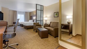 A hotel room with a bed, two sofas, a desk, a chair, a small ottoman, a mirror, and a nightstand with a lamp. - Comfort Suites Little Rock West (Little Rock)