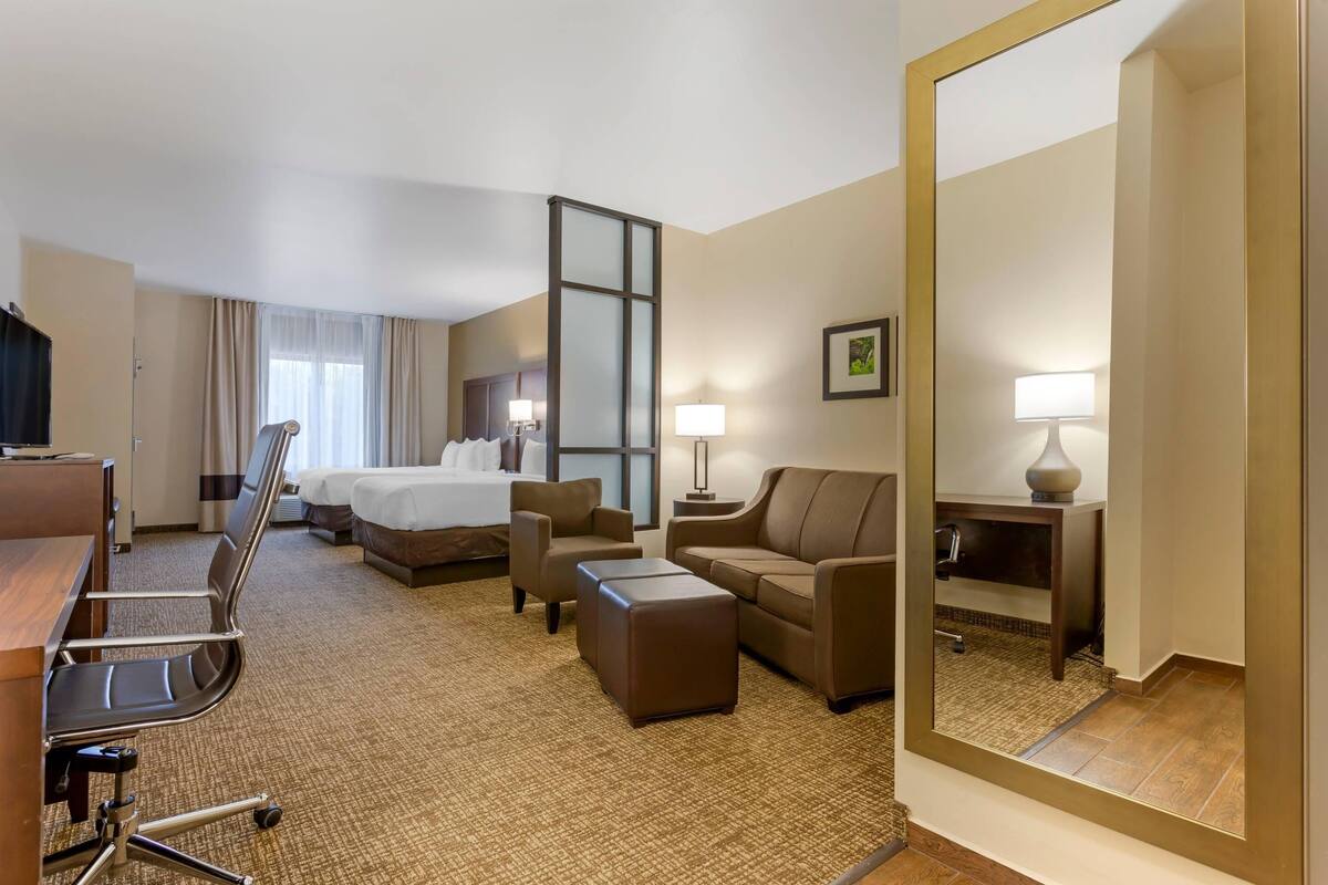 A hotel room with a bed, two sofas, a desk, a chair, a small ottoman, a mirror, and a nightstand with a lamp.