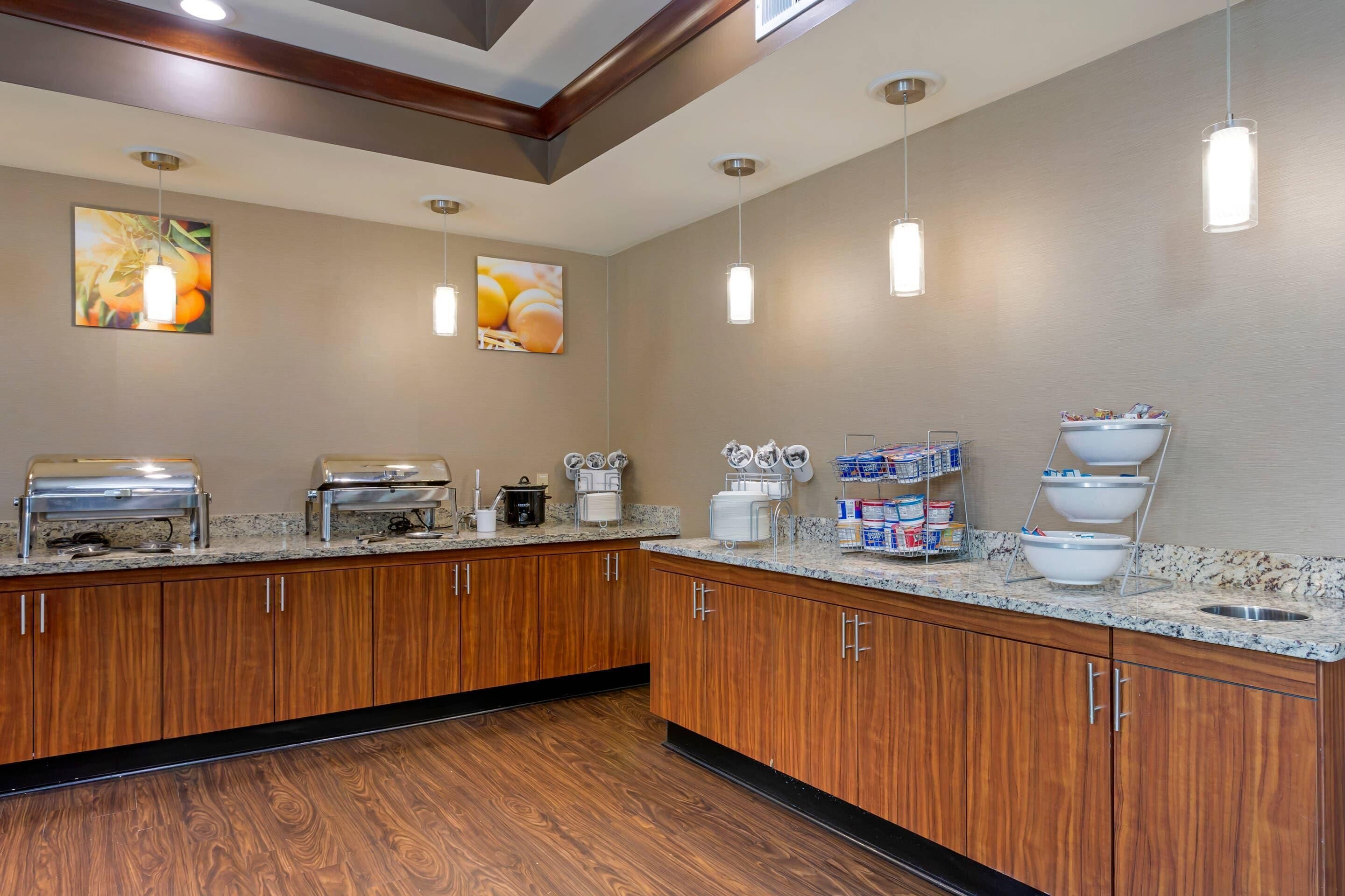 A hotel bathroom with wooden cabinets, a granite countertop, a sink, a coffee maker, and a trash can.