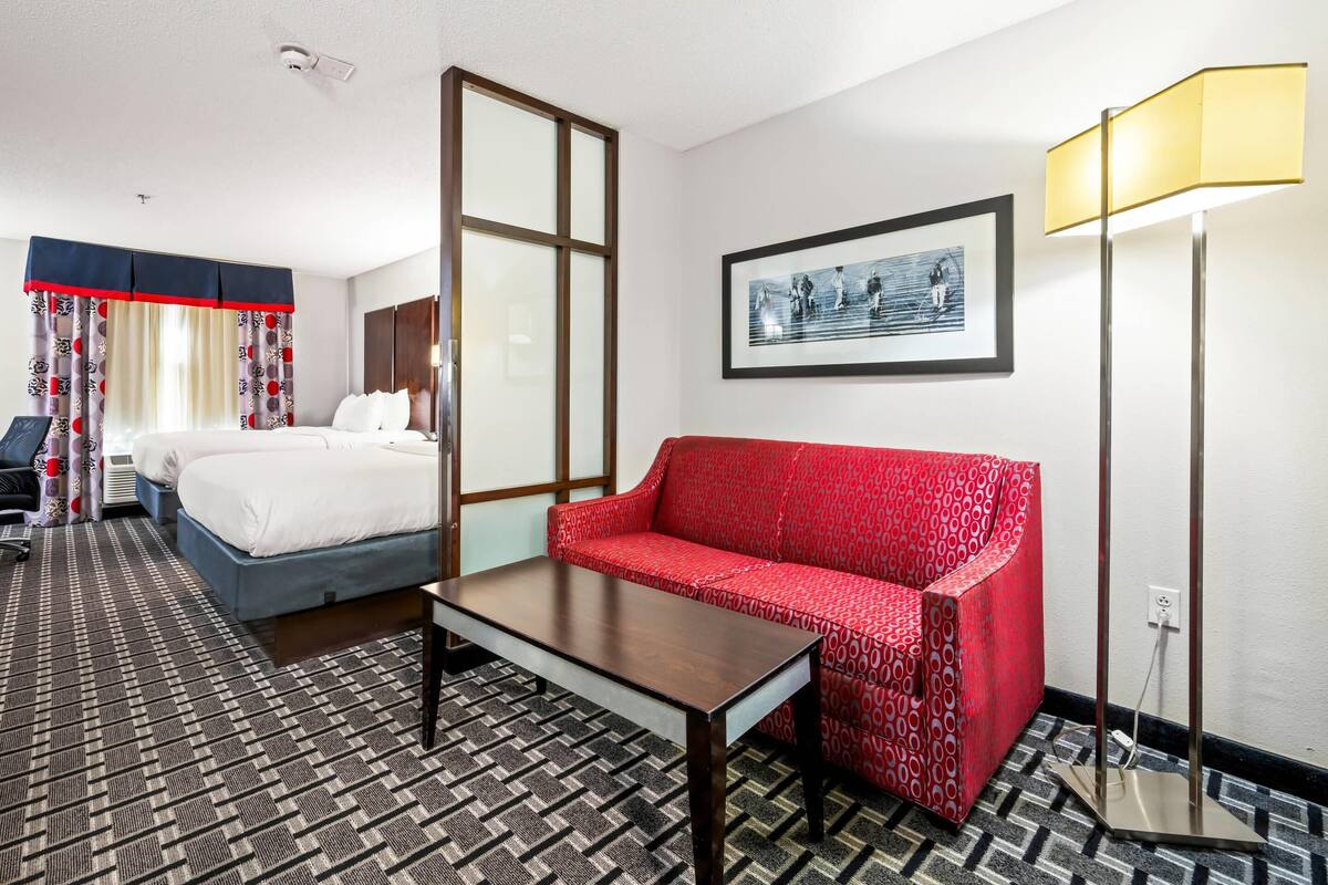 Habitación de hotel con cama, sofá rojo, mesa, lámpara de pie y un cuadro en la pared.