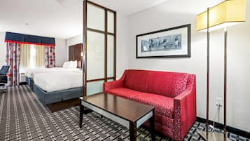 Habitación de hotel con cama, sofá rojo, mesa, lámpara de pie y un cuadro en la pared.