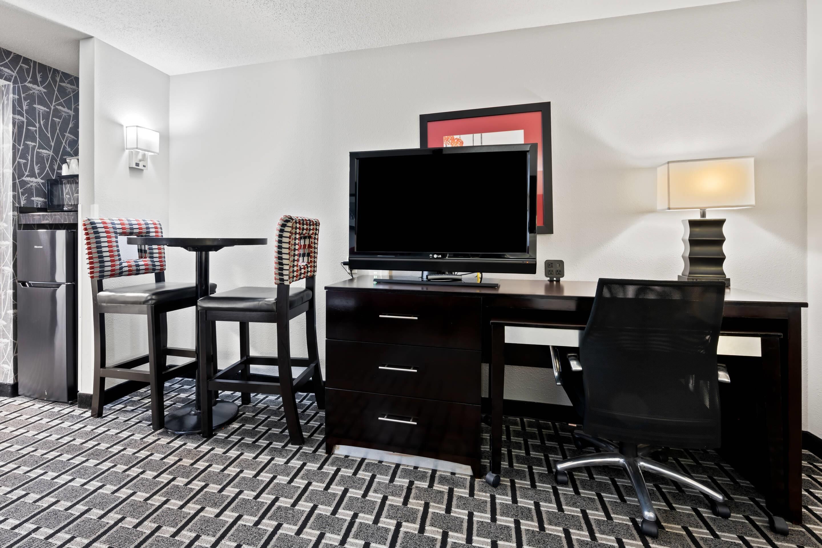 A hotel room with a desk, two chairs, a television, a lamp, and a refrigerator.