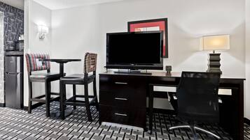 A hotel room with a desk, two chairs, a television, a lamp, and a refrigerator.