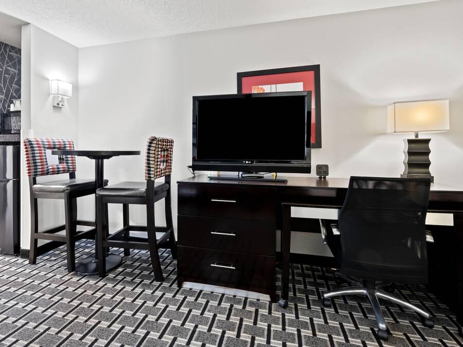 A hotel room with a desk, two chairs, a television, a lamp, and a refrigerator.