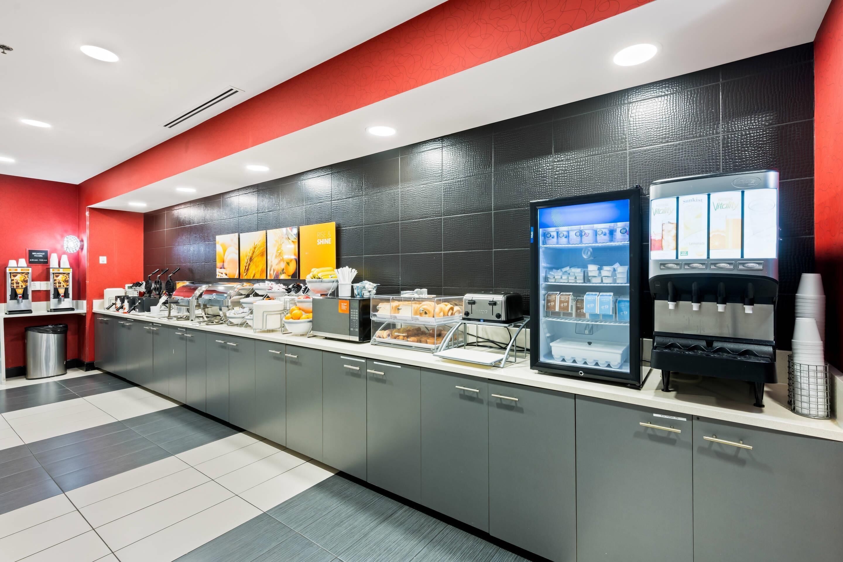 A self-service breakfast area with a coffee machine, a refrigerated drink dispenser, and a variety of food items on the counter.