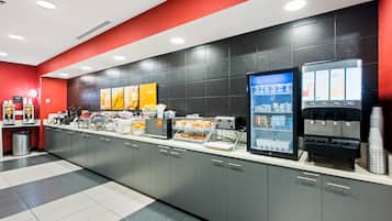 A self-service breakfast area with a coffee machine, a refrigerated drink dispenser, and a variety of food items on the counter.