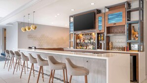 A modern bar area with a long counter, multiple stools, a flat-screen TV, and a variety of bottles and glasses. - Hilton Garden Inn Charlotte/Concord (Concord)