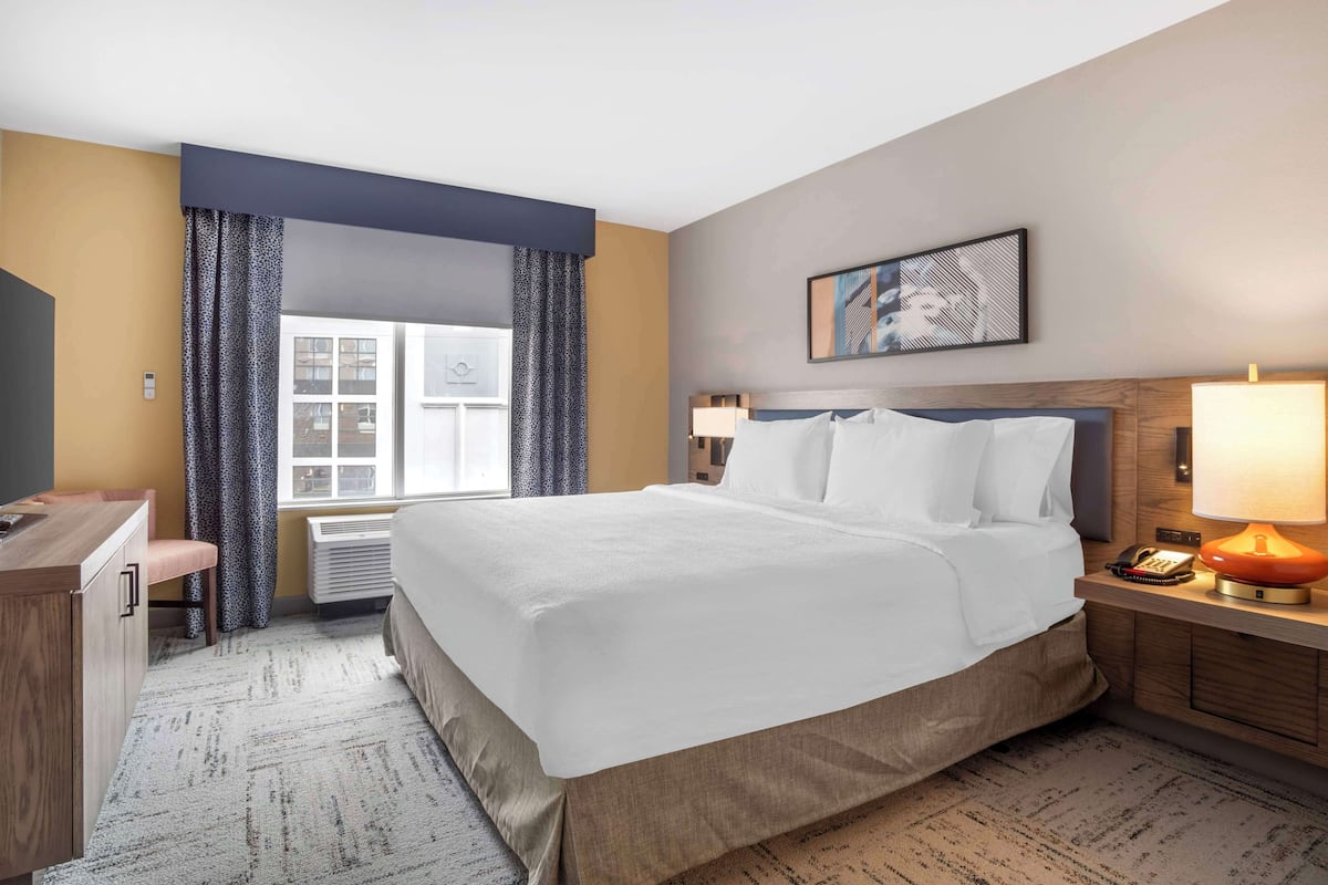 A hotel room with a large bed, a desk, a chair, a window with curtains, a wall-mounted artwork, and a nightstand with a lamp.