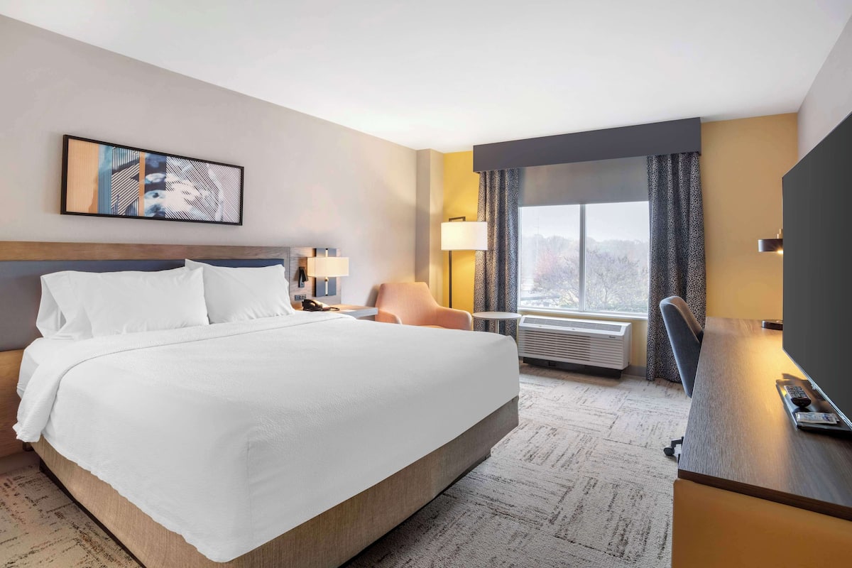 A hotel room with a large bed, a desk, a chair, a window with a view, and a painting on the wall.