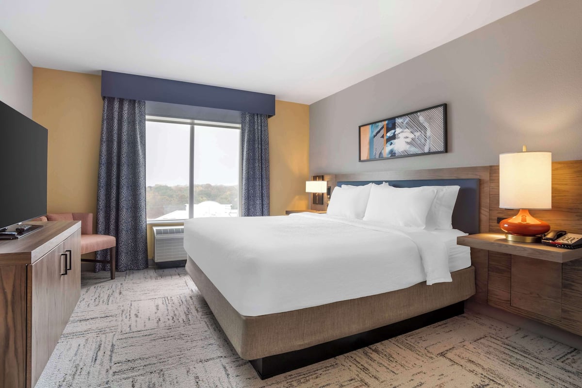 A hotel room with a large bed, a desk, a chair, a window with curtains, a wall-mounted TV, and a painting.