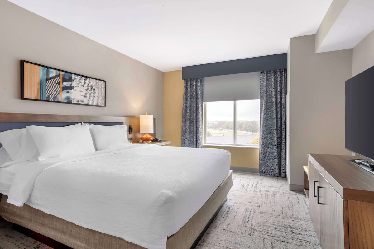 A hotel room with a large bed, a desk with a TV, a window with curtains, and a painting on the wall.