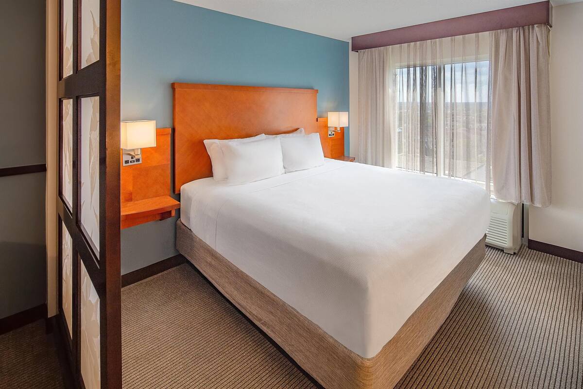 A hotel room with a large bed, wooden headboard, two bedside lamps, and a view of the city through sheer curtains.