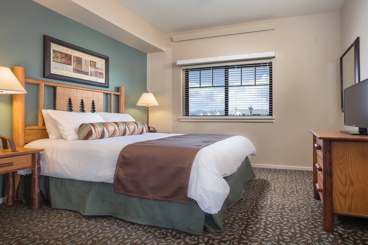 A hotel room with a bed, two bedside tables with lamps, a television, a window with blinds, and a framed picture on the wall.
