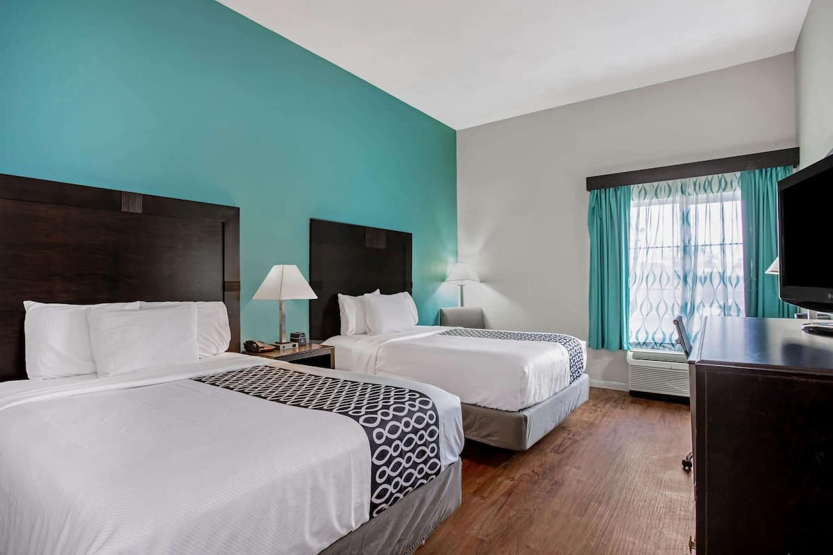 Une chambre d’hôtel avec deux lits, une tête de lit en bois foncé, des murs de couleur turquoise et une fenêtre ornée de rideaux turquoise.