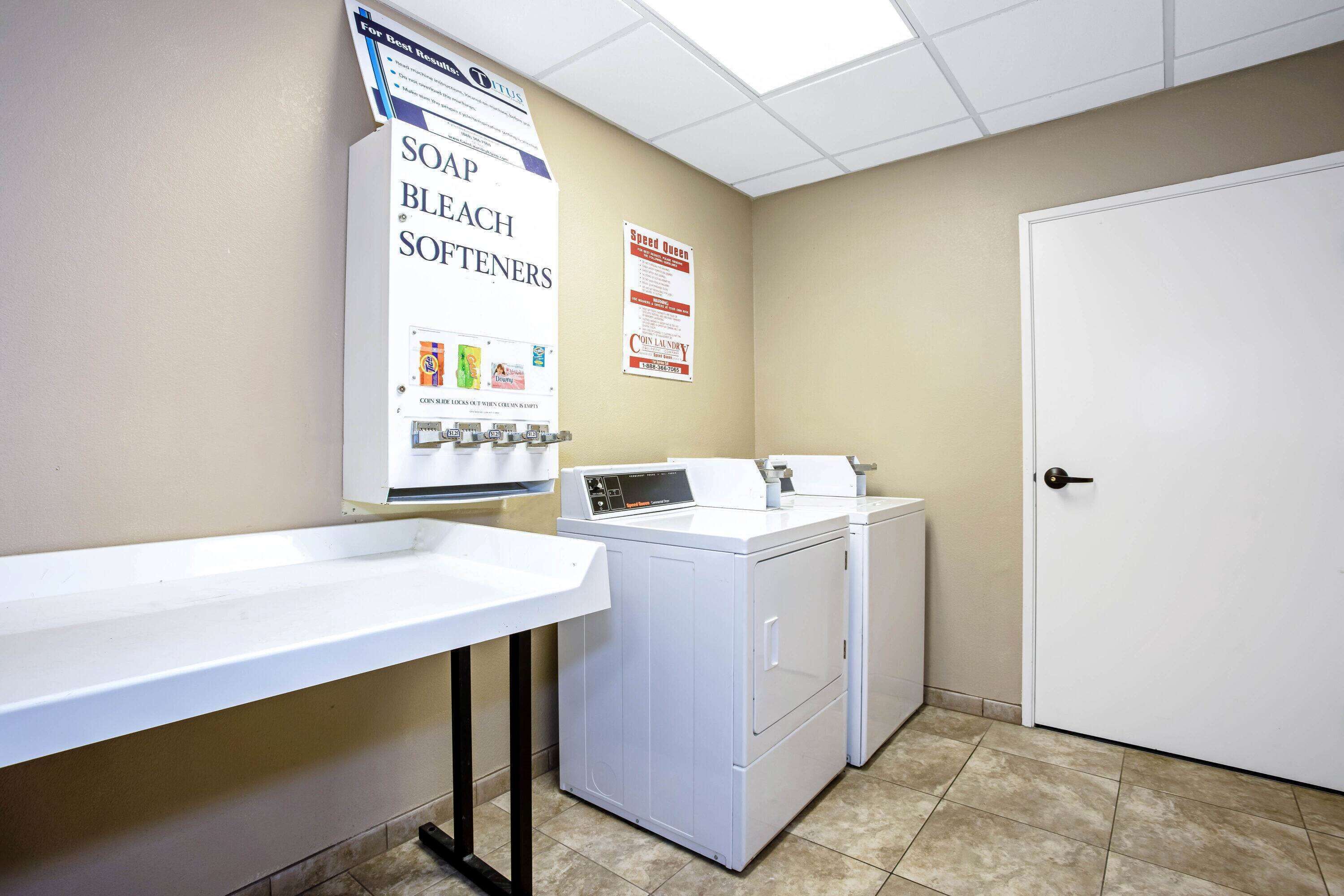 A laundry area with a sink, a washing machine, and a dryer. There are signs for soap, bleach, and softeners.