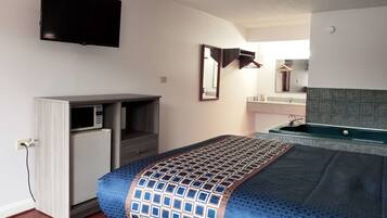 A compact hotel room with a bed, a built-in TV, a small kitchenette, and a bathroom with a mirror and sink.