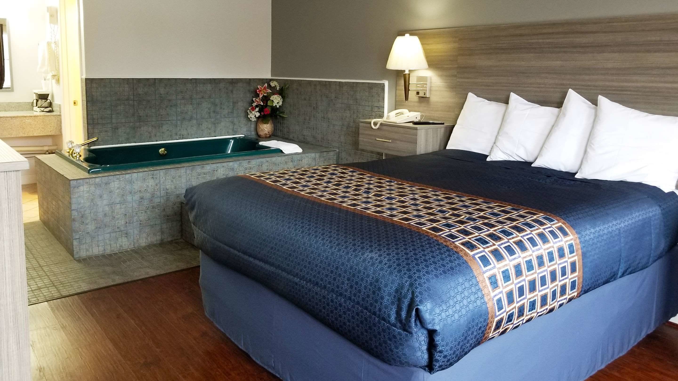 Et hotelværelse med en stor seng, et lille badeværelse med badekar, et natbord med lampe og en vase med blomster.