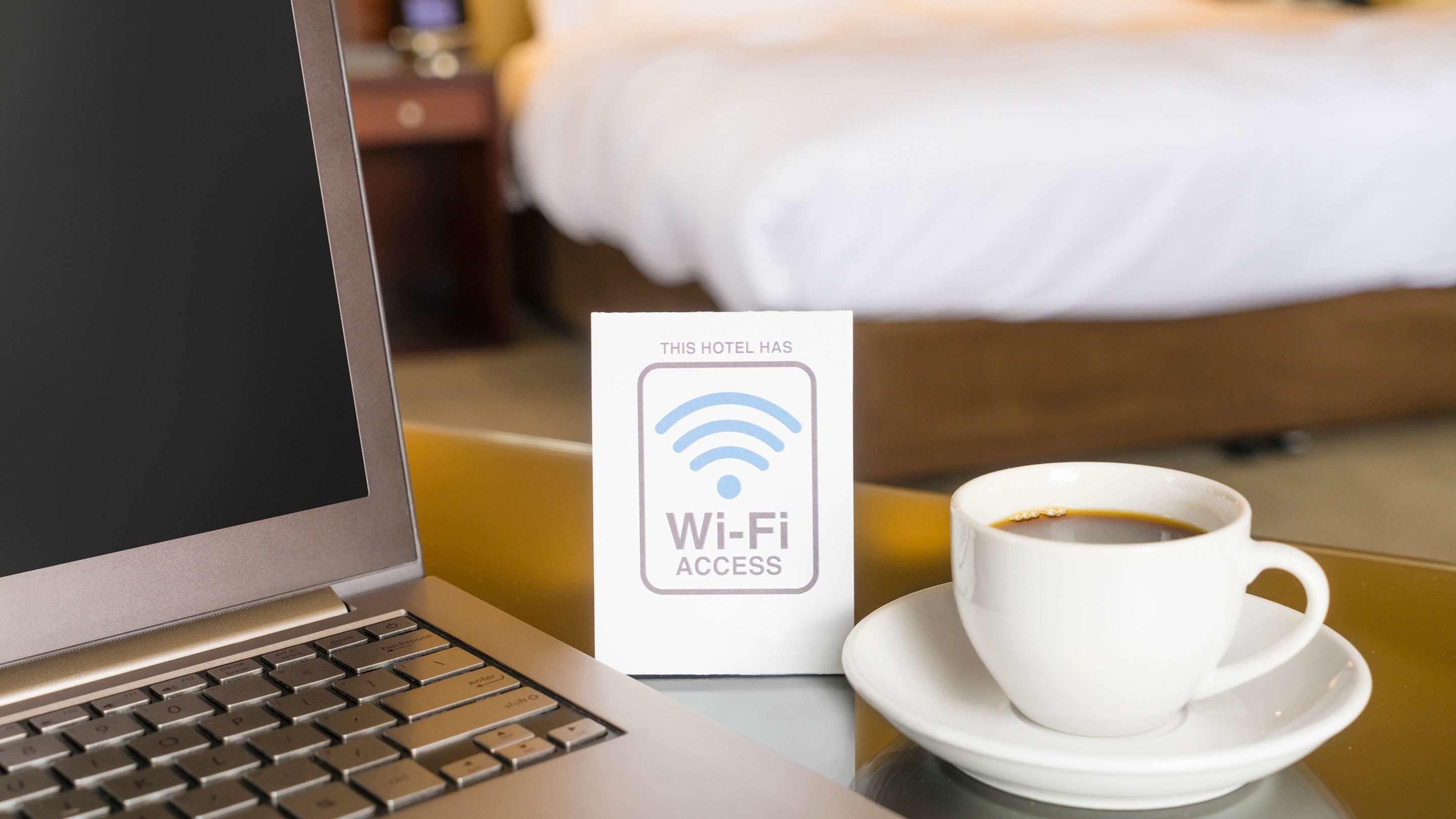 A laptop, a Wi-Fi access sign, and a cup of coffee on a table in a hotel room.
