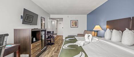 Una habitación de hotel con una cama, un escritorio con una silla, un aparador, un televisor montado en la pared y un cuadro enmarcado.