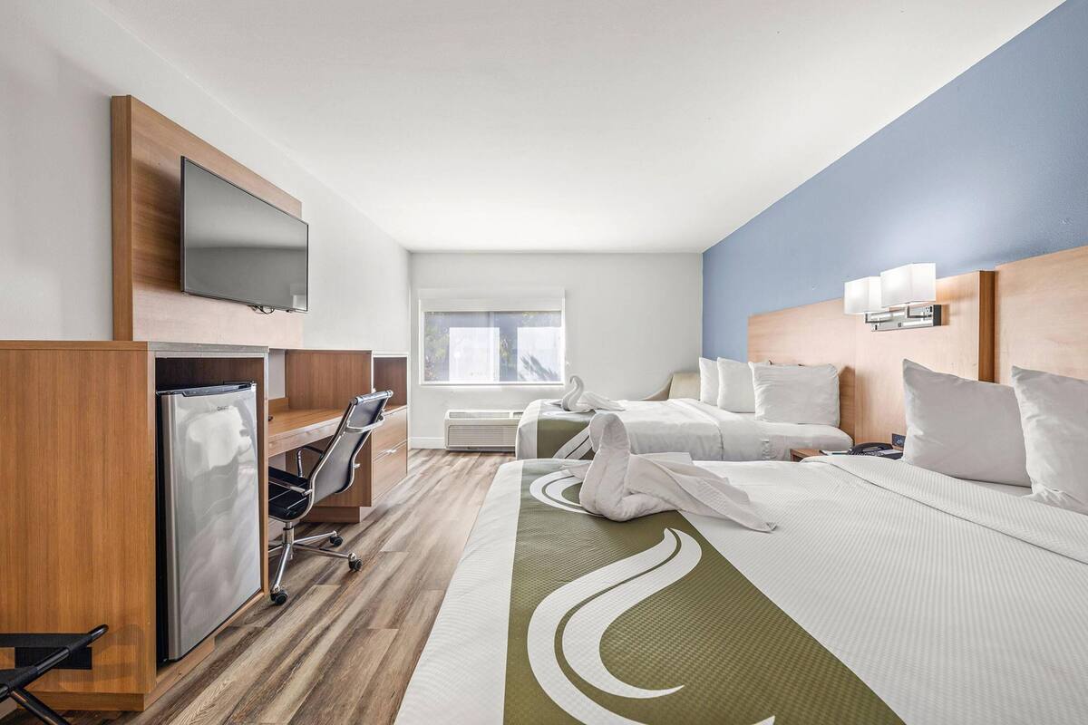 A hotel room with a bed, a desk with a computer, a refrigerator, a TV mounted on the wall, and a window with a view of the outdoors.