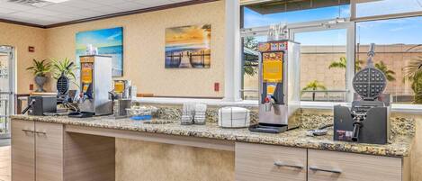 A kitchen area with a granite countertop, coffee machines, waffle maker, and a view of the outside.