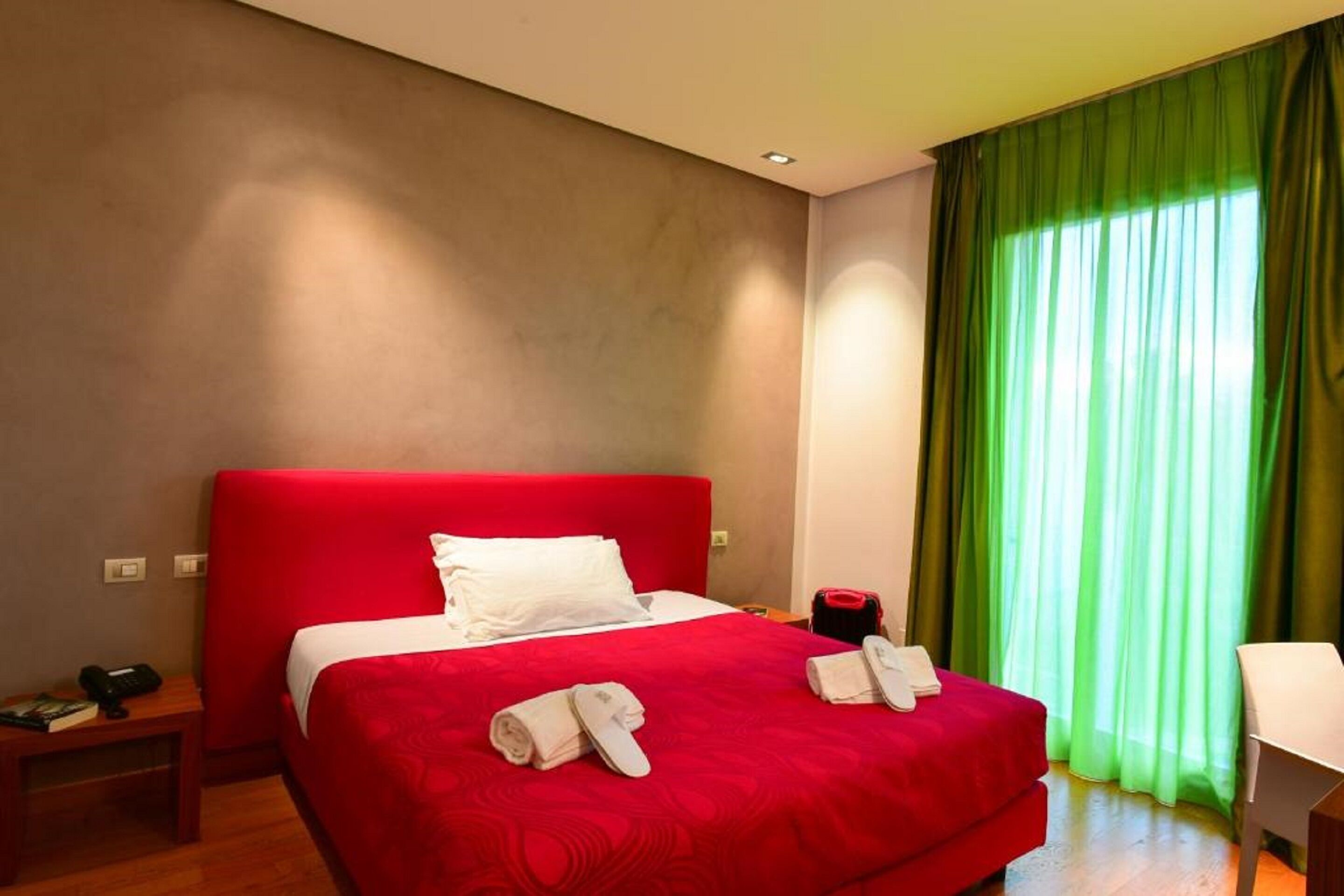 A hotel room with a red bed, green curtains, a white desk, and a wooden floor.