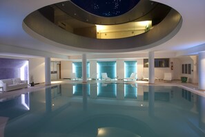 A modern indoor swimming pool with a central skylight and lounge chairs around the perimeter.