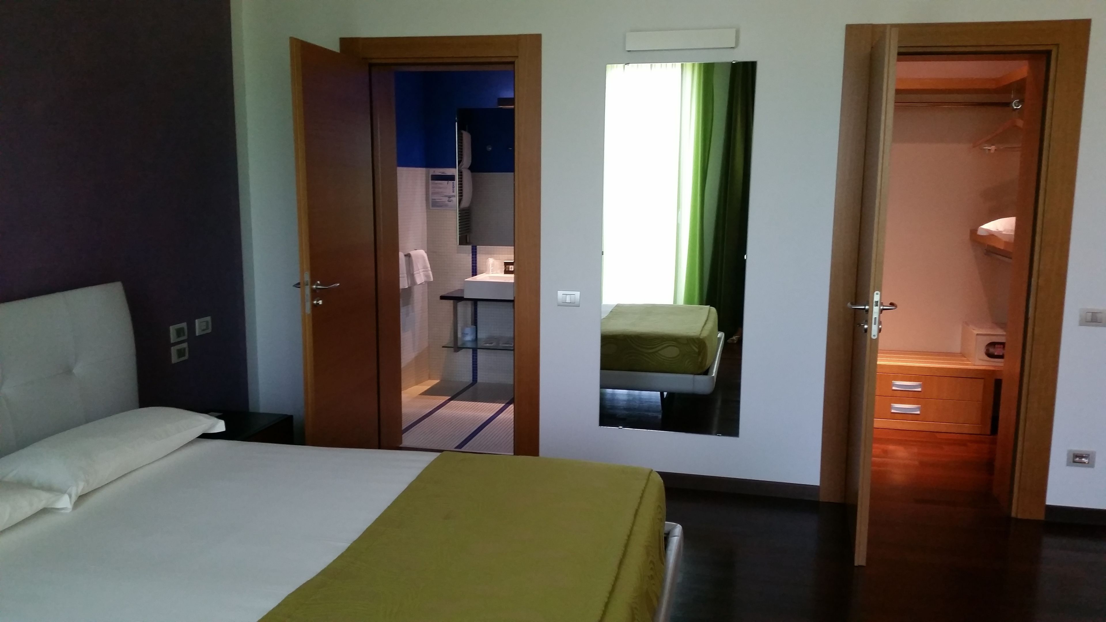 Hotellihuone, jossa on sänky, avoimesta ovesta näkyvä kylpyhuone ja pieni oleskelualue, jossa on tuoli.
