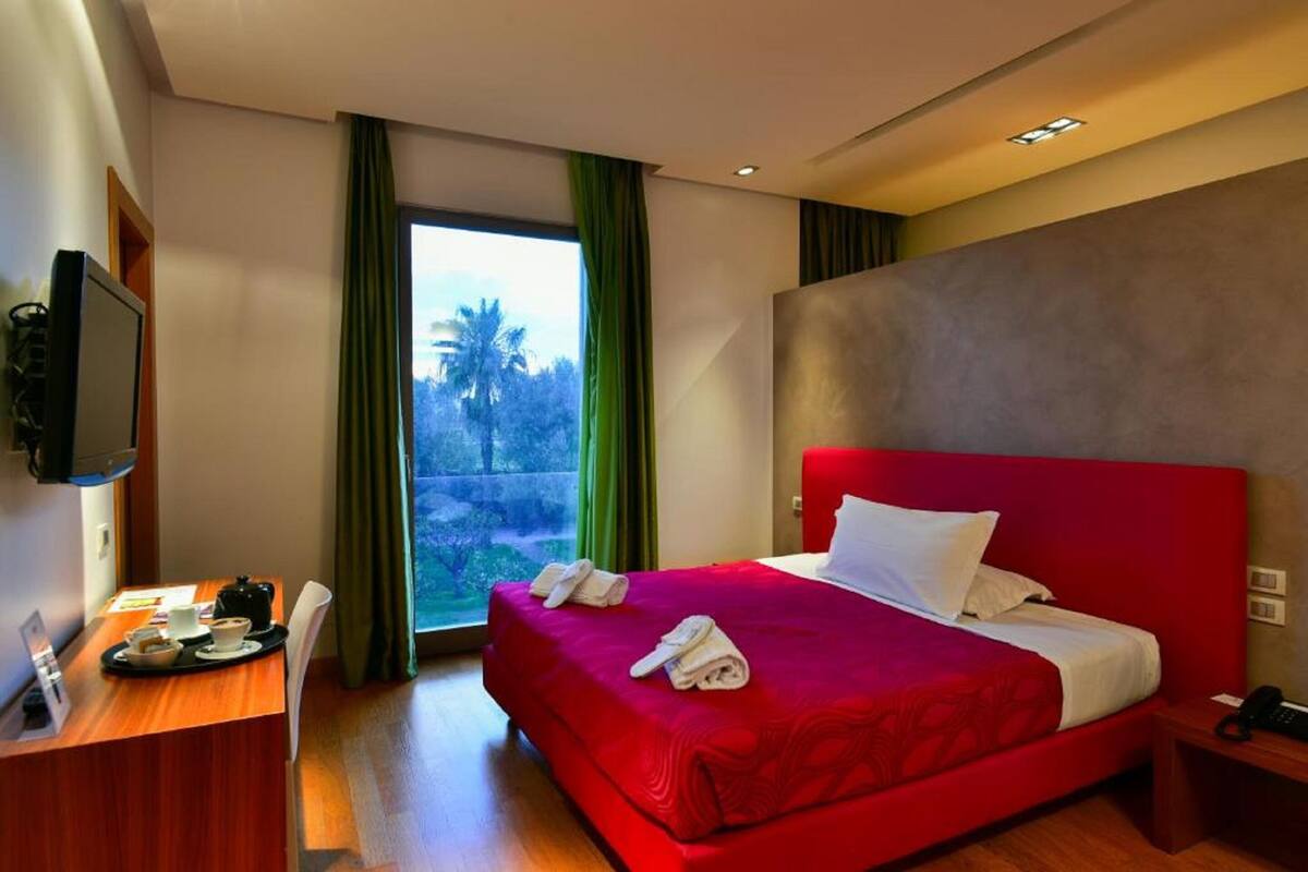 A hotel room with a large bed, a desk with a chair, a TV mounted on the wall, and a window with curtains.