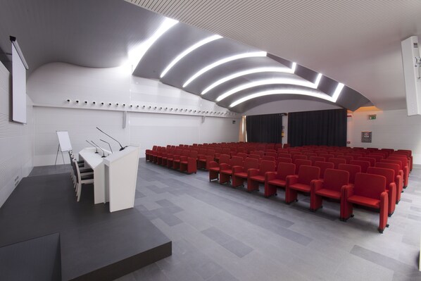 A modern auditorium with rows of red chairs, a podium, and a curved ceiling with integrated lighting.