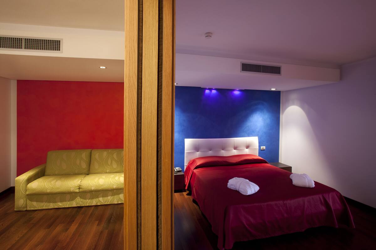 A hotel room with a red and blue accent wall, a green sofa, a bed with red bedding, and a wooden door frame.