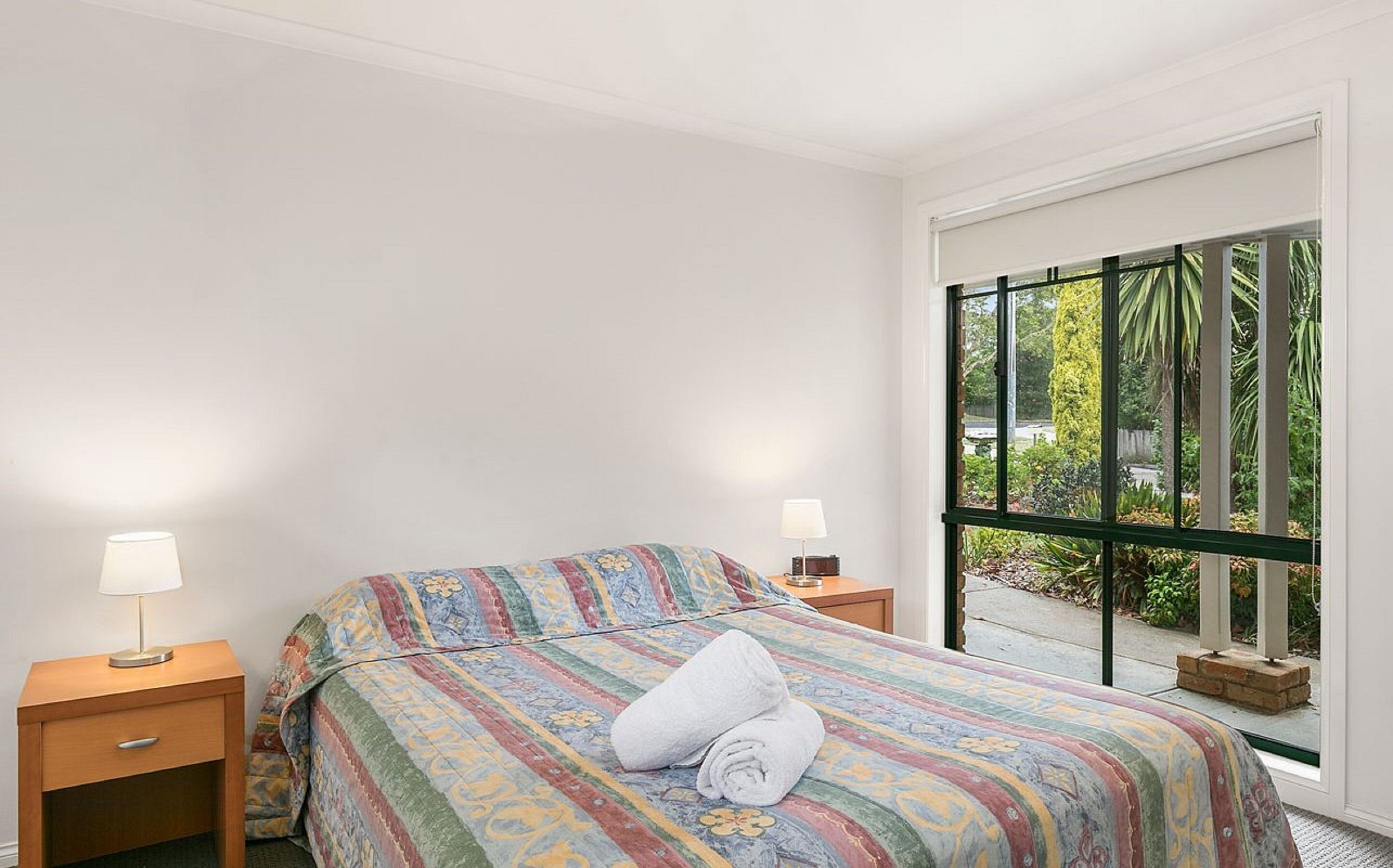 A bedroom with a bed, two bedside tables, a window with a view of greenery, and a white wall.