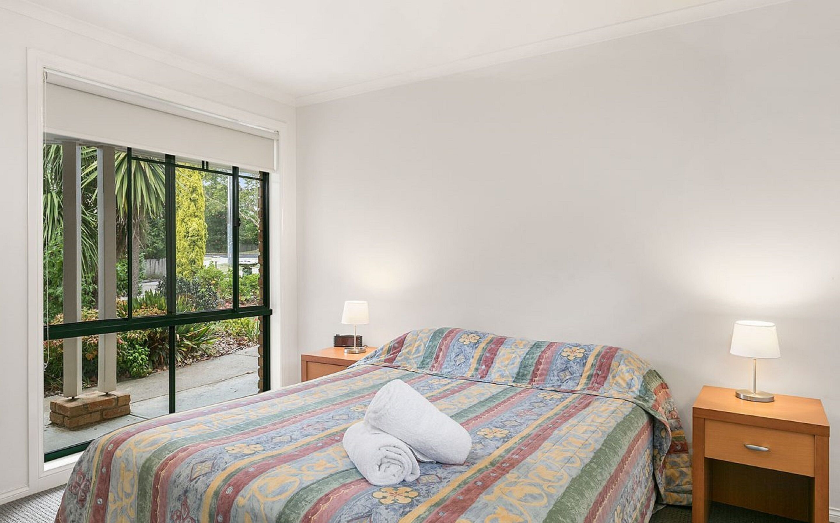 A bedroom with a bed, two bedside tables, and a window with a view of greenery.
