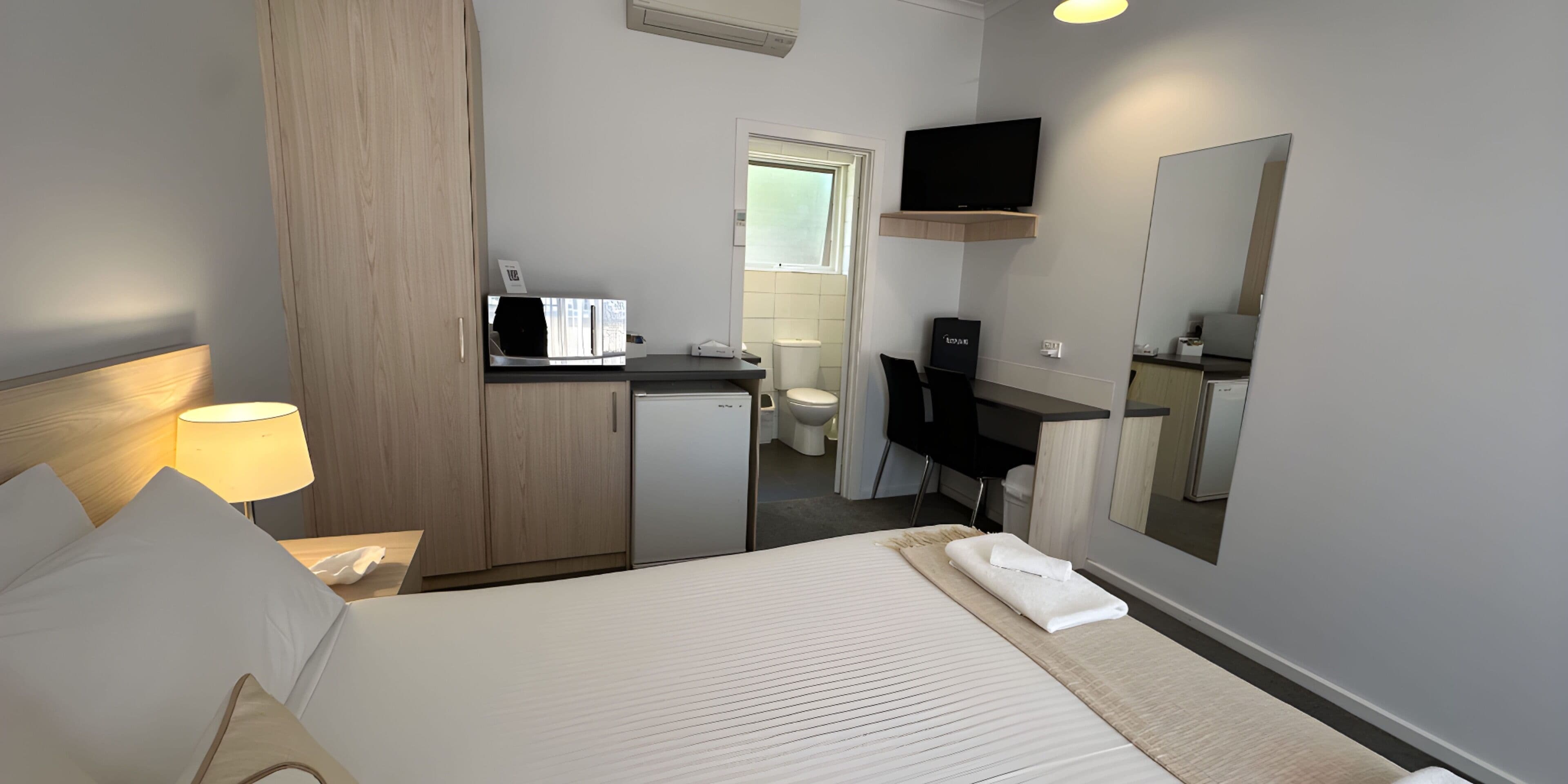 A hotel room with a bed, a desk, a television, a mirror, and a small kitchenette.