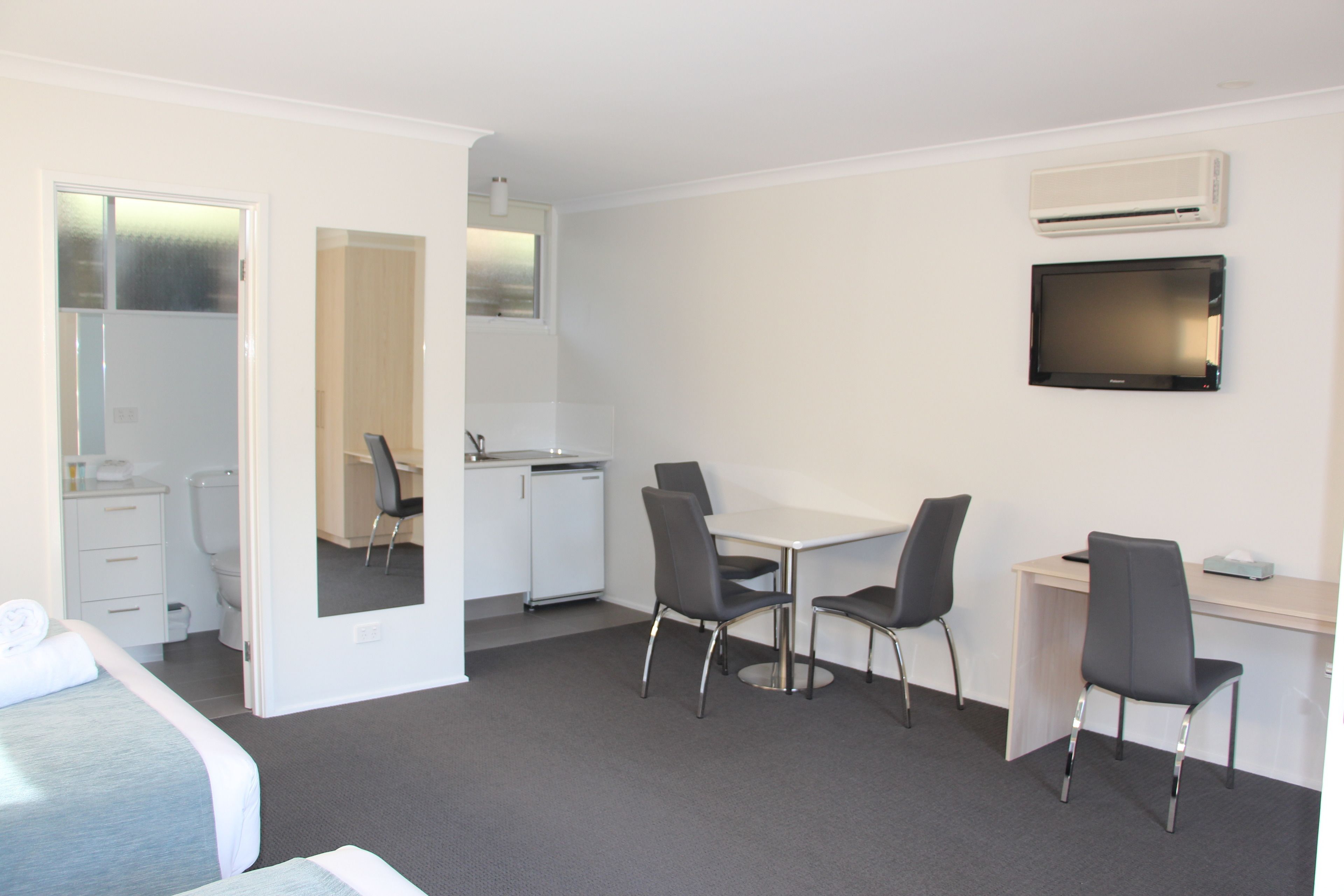 A hotel room with a bed, a desk, two chairs, a small kitchenette, and a TV mounted on the wall.
