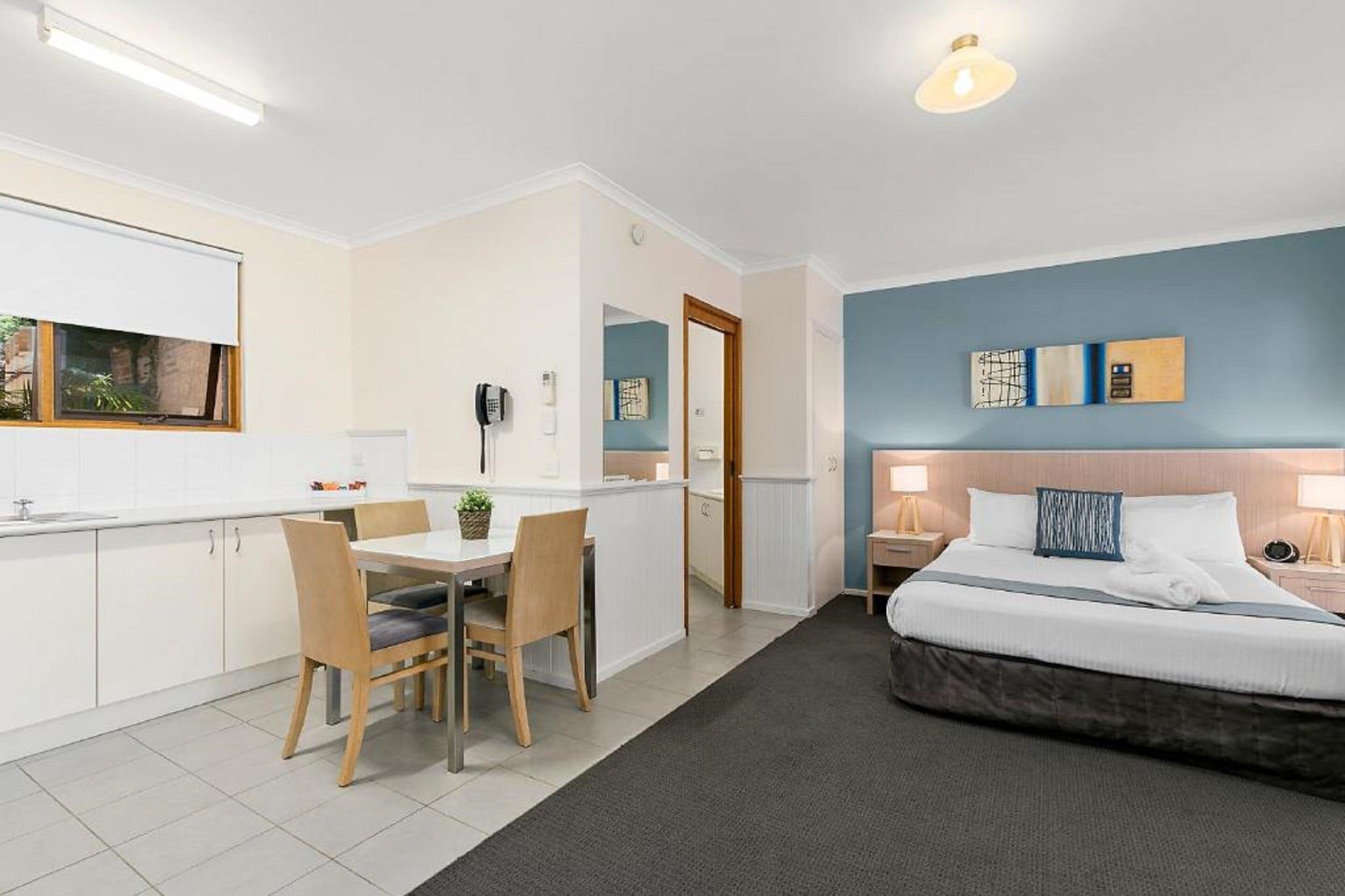 A modern hotel room with a bed, bedside tables, a desk with chairs, a small kitchen area, and a bathroom.