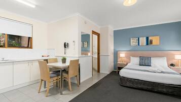 A modern hotel room with a bed, bedside tables, a desk with chairs, a small kitchen area, and a bathroom.