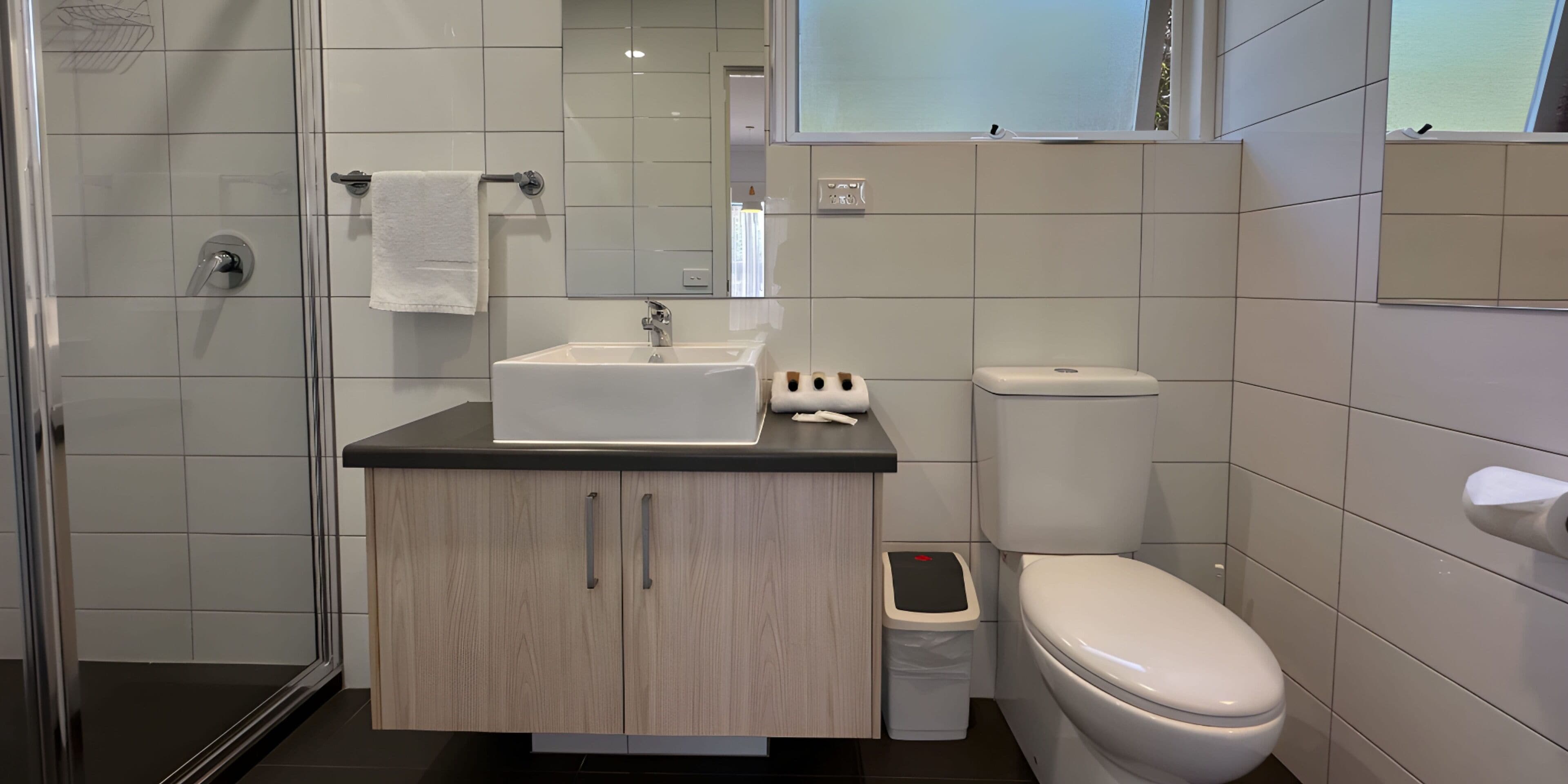 Een moderne badkamer met een douchecabine van glas, een witte wastafel met een zwart aanrecht, een toilet en een kleine prullenbak.