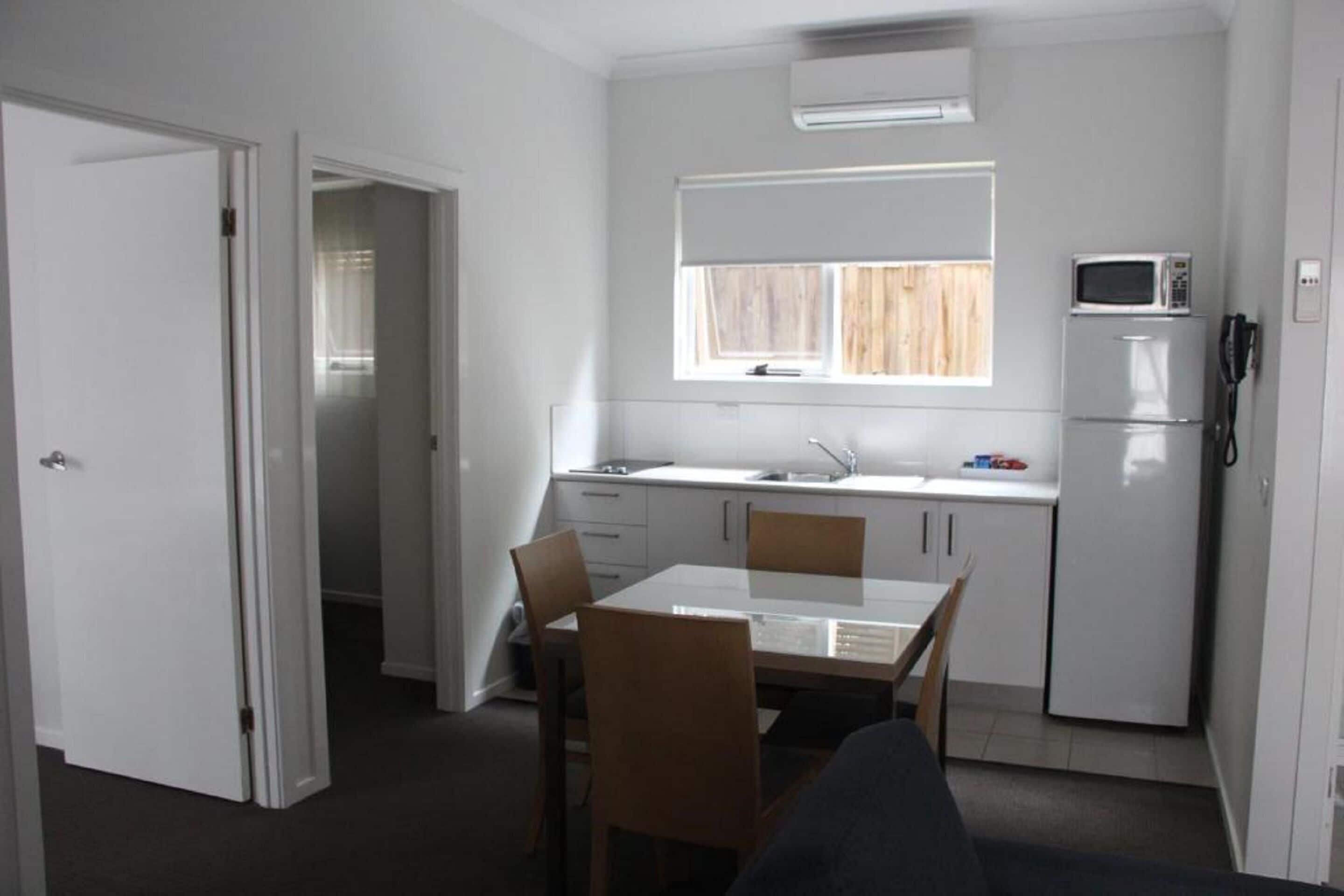 Cocina compacta con muebles blancos, microondas y refrigerador. Hay una ventana sobre el fregadero y una puerta que da a otra habitación.