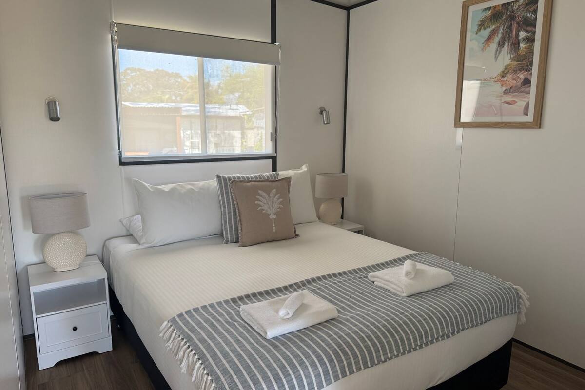 A neatly arranged bedroom with a large bed, a bedside table with a lamp, a window with a view, and a framed picture on the wall.
