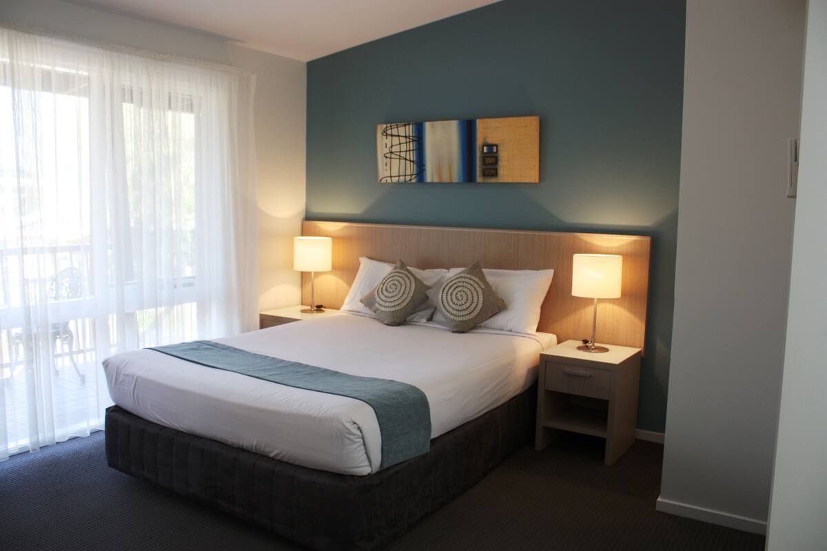 A hotel room with a large bed, two bedside tables with lamps, a view of the outdoors through sheer curtains, and two wall-mounted artworks.
