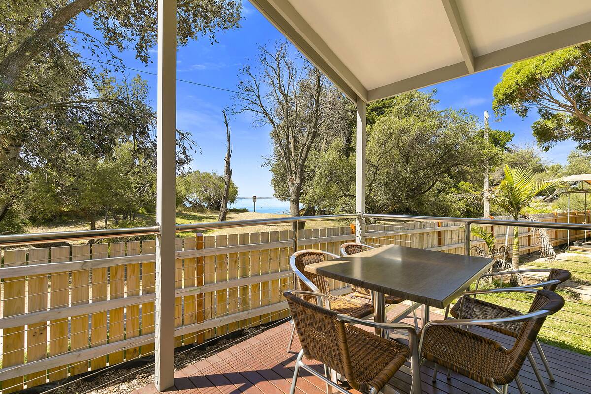 A covered outdoor patio with a table and wicker chairs, overlooking a fenced area with trees and a body of water.