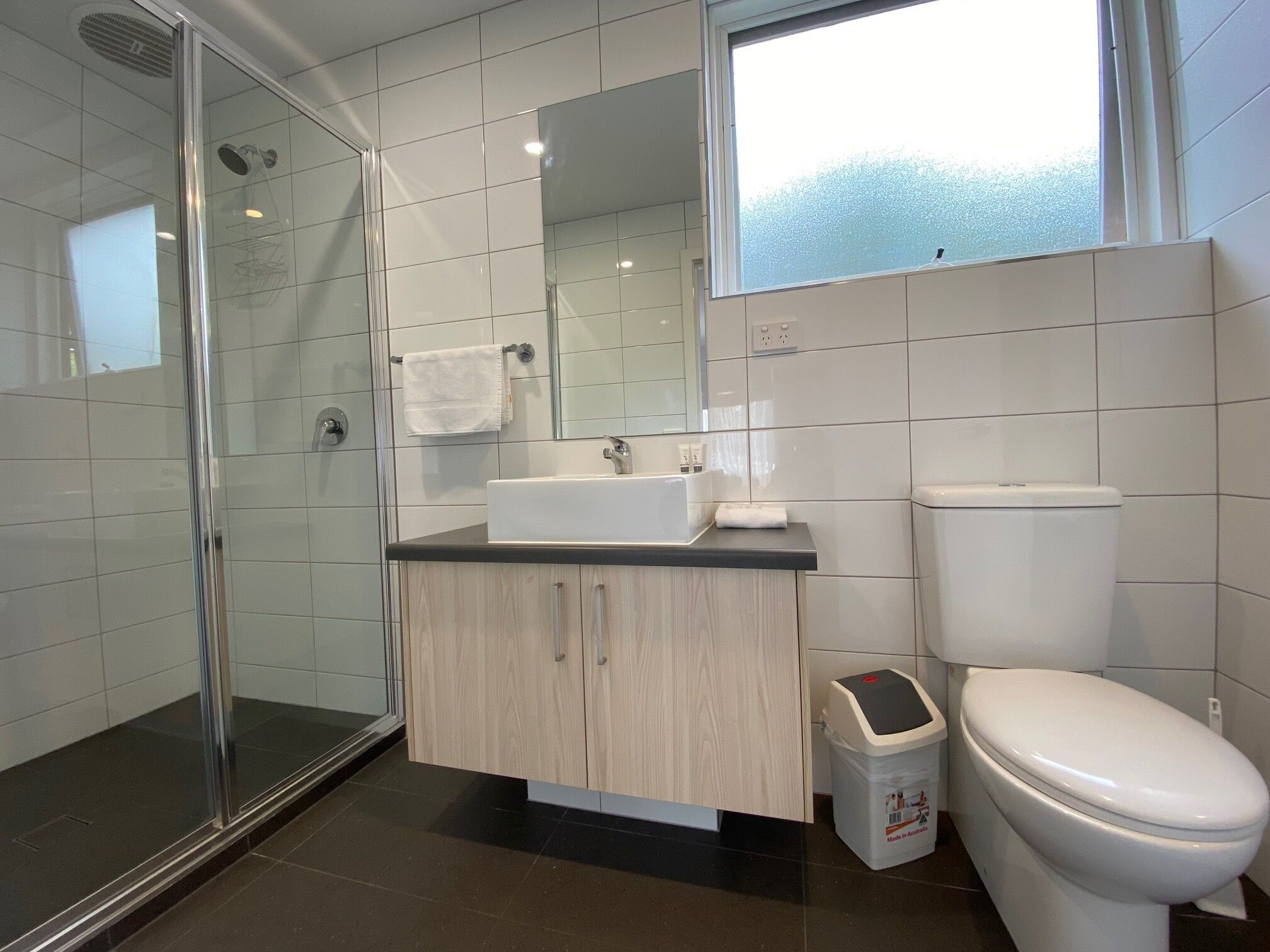A modern bathroom with a glass shower enclosure, a white toilet, a sink with a mirror above it, and a small trash bin.