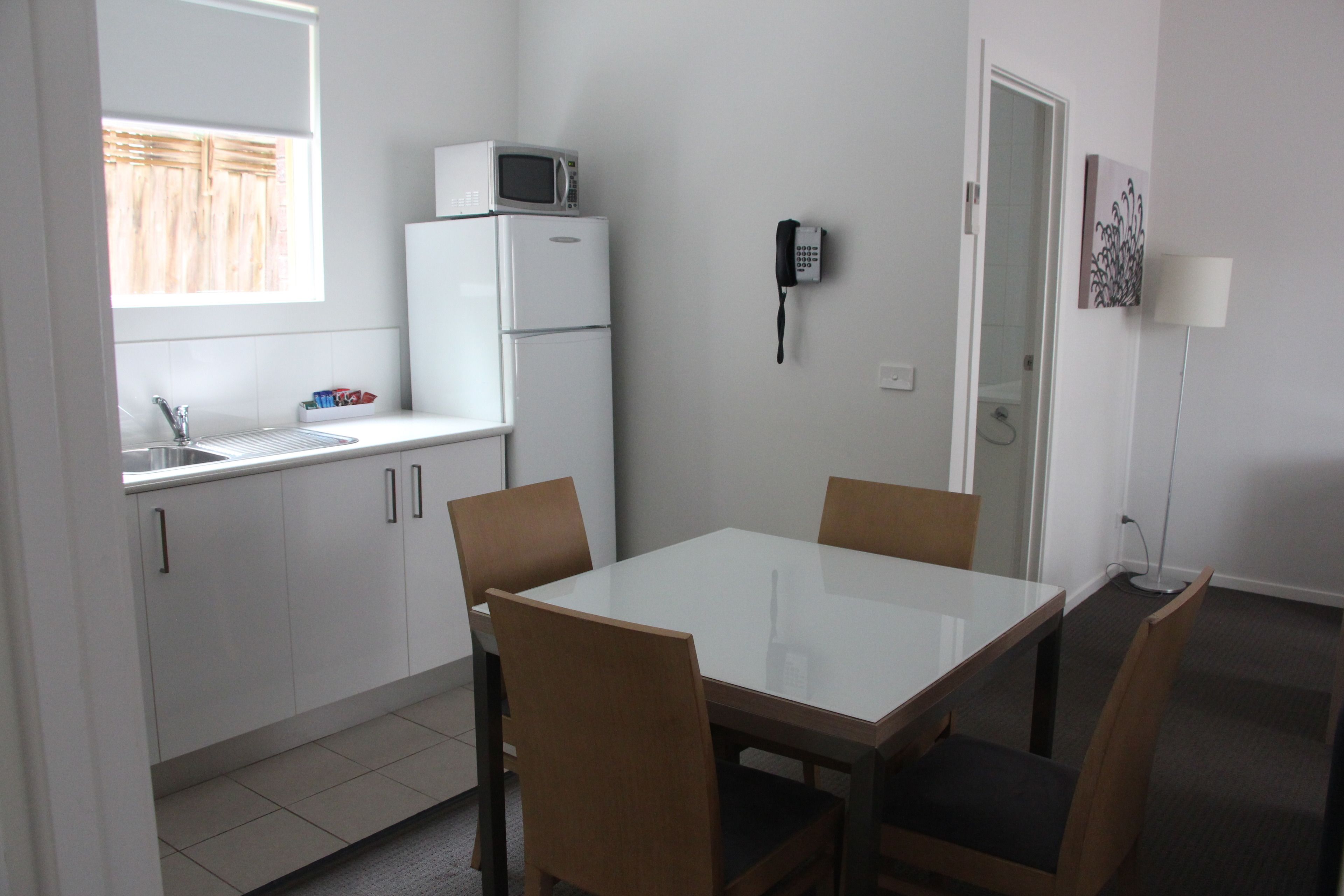Cocina compacta con microondas, refrigerador y teléfono en la pared. Hay una mesa de comedor con sillas y una ventana con persianas.
